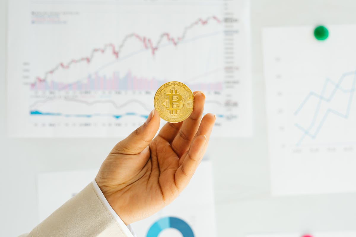 Person holding a Bitcoin in front of financial charts displayed on a board.