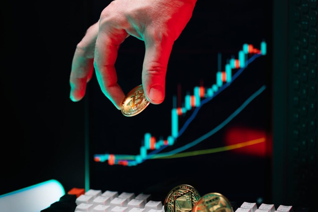 Hand holding a Bitcoin with stock market charts on a computer screen in the background.