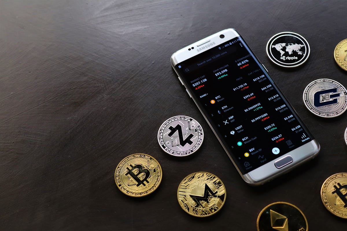 Smartphone displaying cryptocurrency values surrounded by various physical crypto coins on a dark surface.