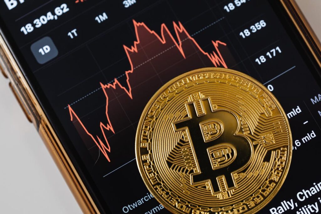 A Bitcoin coin in front of a smartphone displaying cryptocurrency market trends with red graphs.