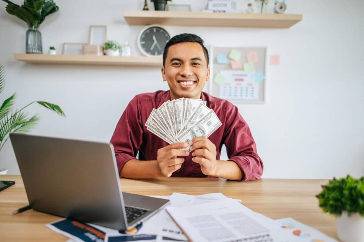 A person in a maroon shirt holds a fan of cash in front of a laptop