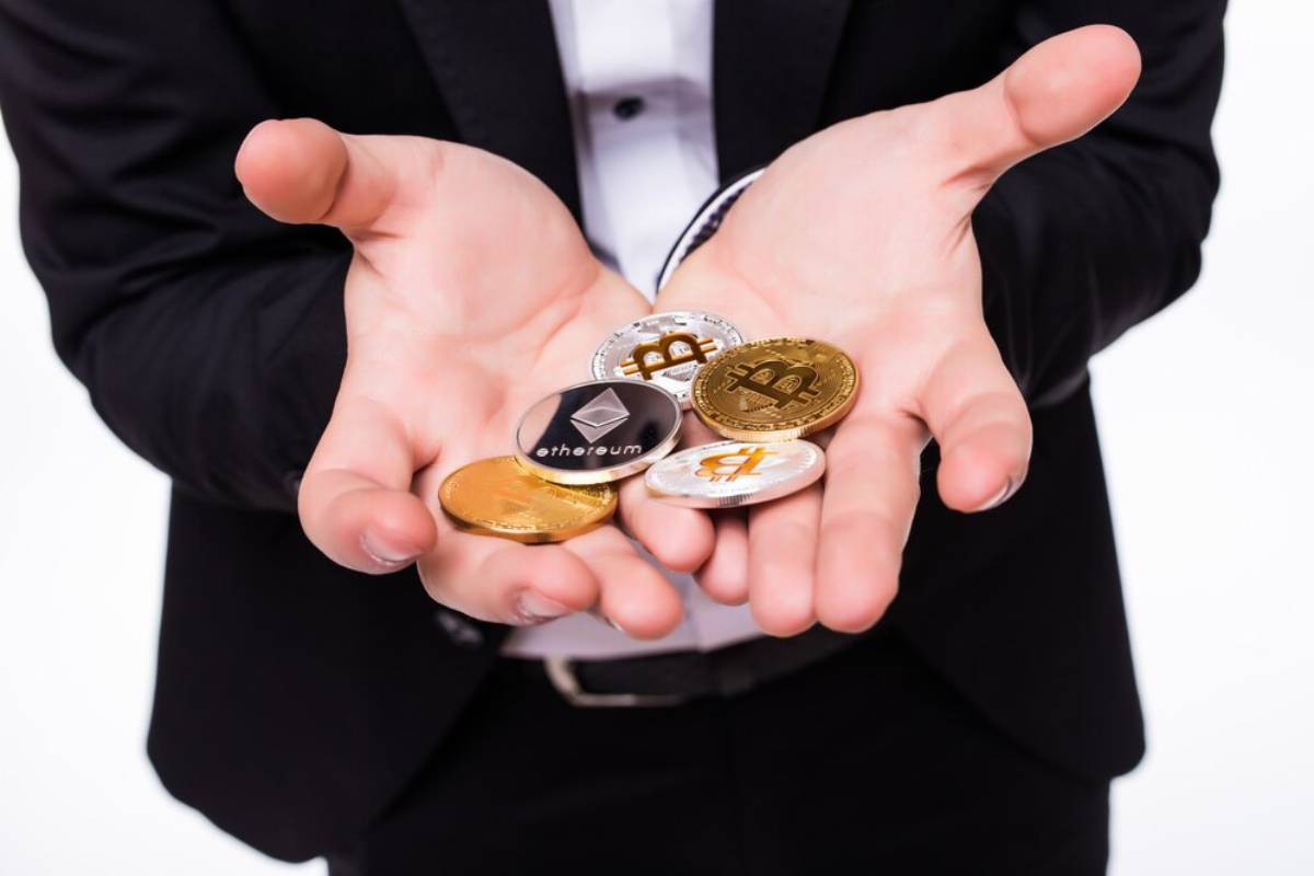 Person in a suit offering various cryptocurrency coins including Bitcoin and Ethereum held in open hands, against a white background.