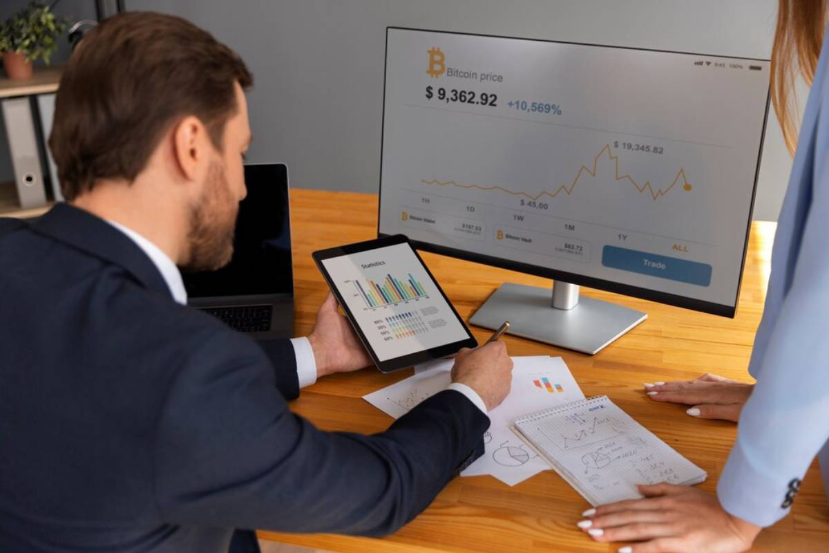 Two professionals examine financial data on a tablet and monitor displaying Bitcoin prices in an office.