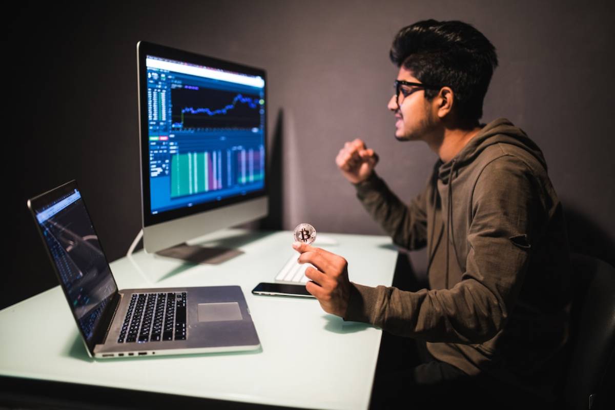 Young man analyzing financial charts on computer screens, holding a physical Bitcoin, in a dark room.