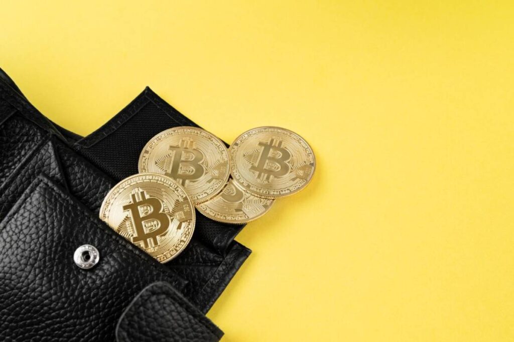 Three Bitcoin coins spilling from a black leather wallet on a yellow background.