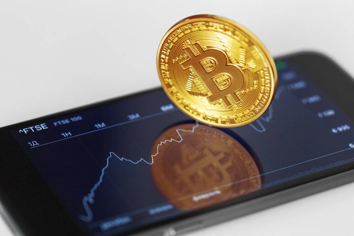 A Bitcoin coin balances on a smartphone displaying stock market graphs.