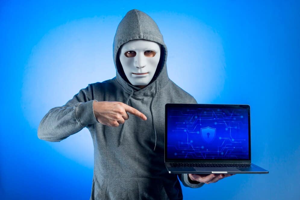 Person in mask and hoodie pointing at a laptop screen displaying a cybersecurity shield symbol on a blue background.
