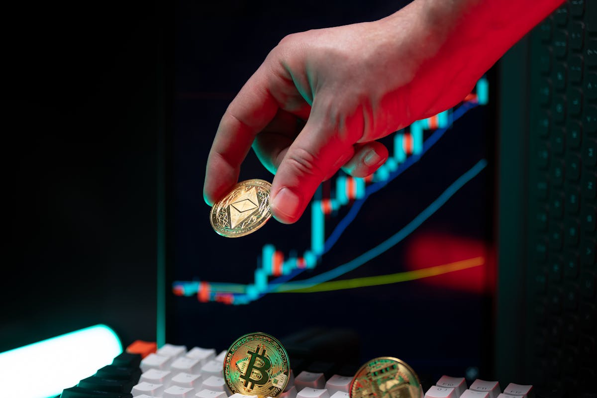 A hand holding a cryptocurrency coin over a keyboard with financial graphs on a screen in the background.