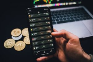 Hand holding a smartphone displaying cryptocurrency prices, with physical coins and a laptop in the background.