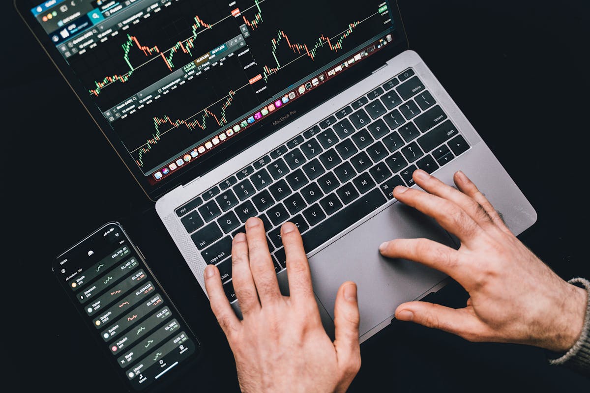 Hands navigating a laptop and smartphone displaying stock market charts, emphasizing active trading or analysis.