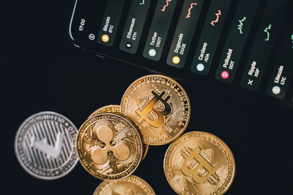 Various cryptocurrency coins including Bitcoin and others in front of a smartphone screen showing live trading graphs.