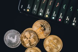 Various cryptocurrency coins including Bitcoin and others in front of a smartphone screen showing live trading graphs.