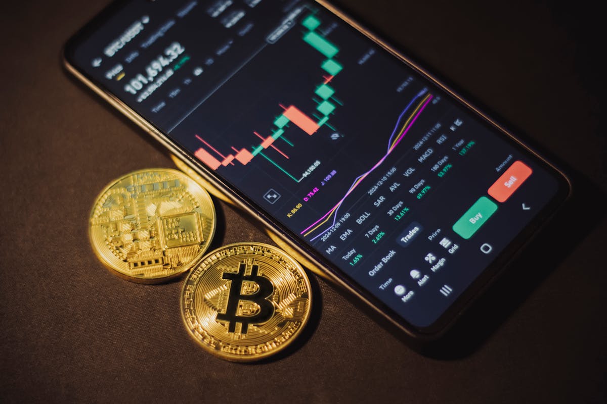 Smartphone displaying cryptocurrency trading application with Bitcoin tokens nearby on a dark surface.