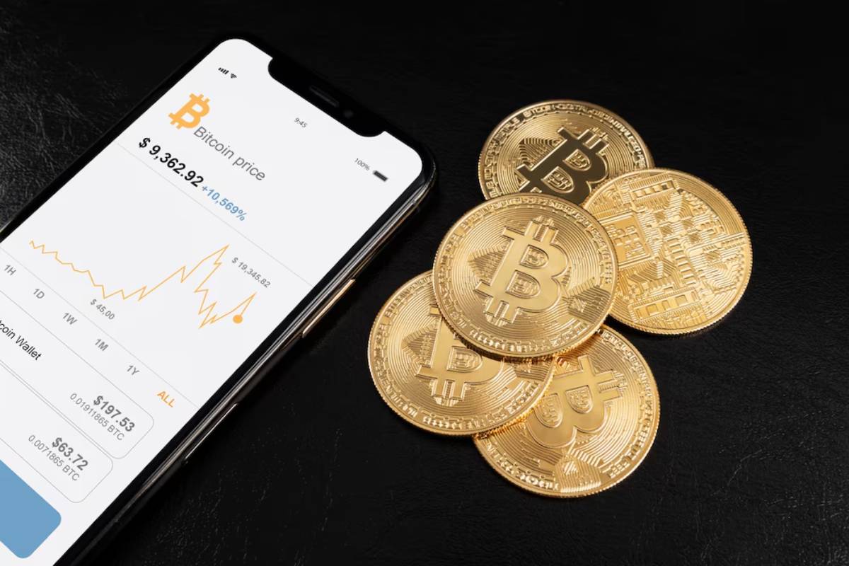 Smartphone showing Bitcoin price chart beside physical Bitcoin tokens on a dark surface.