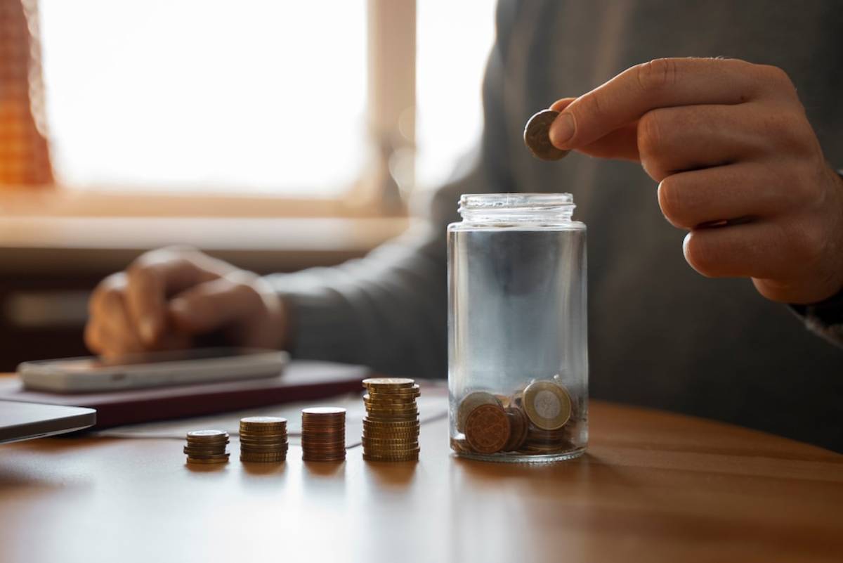 A person drops a coin into a clear jar, surrounded by stacks of coins