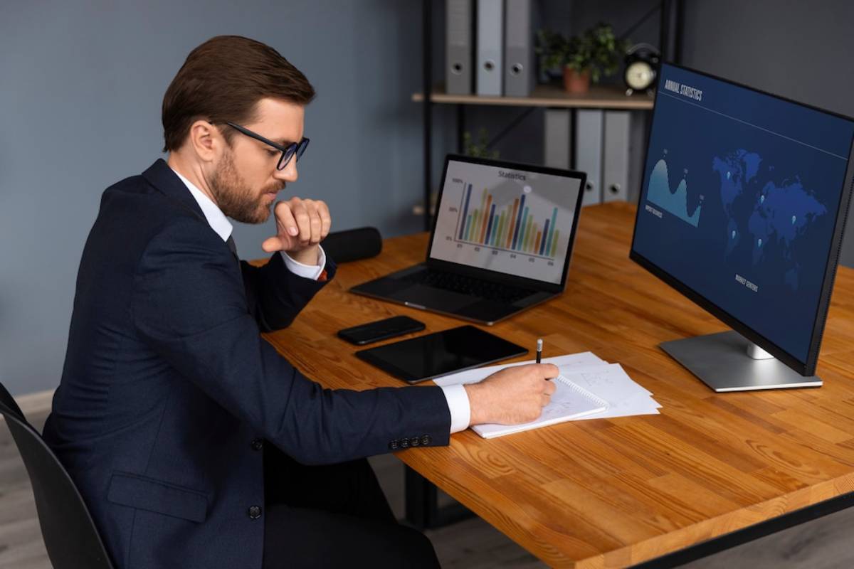A person in a suit sits at a wooden desk, noting data while looking at graphs