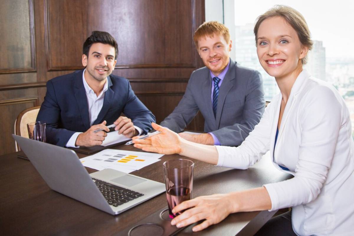 A business meeting in a modern conference room, with individuals engaged in discussion around a laptop