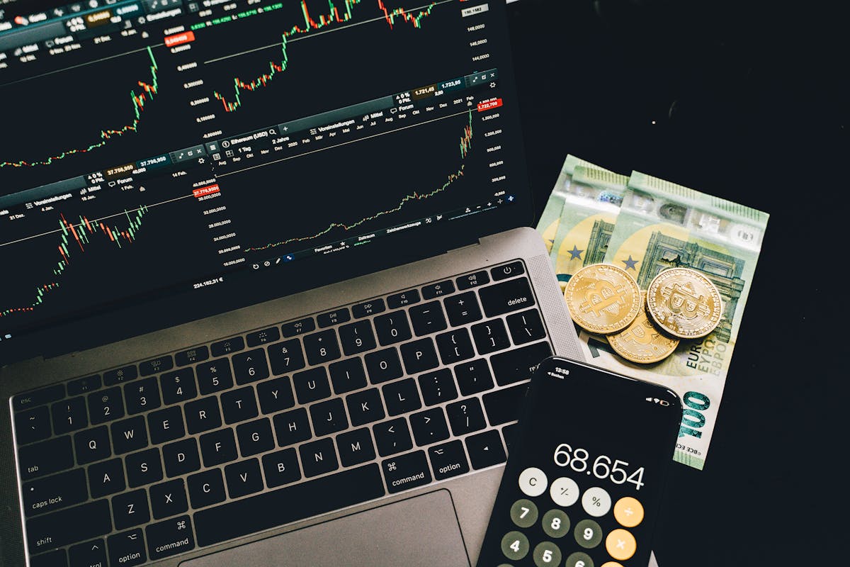 Laptop displaying stock charts, Bitcoin coins, Euro bills, and a calculator on a dark desk.