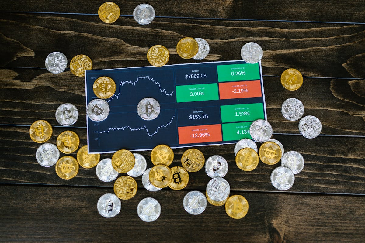 A digital tablet displaying cryptocurrency market data surrounded by various gold and silver crypto coins on a wooden surface.