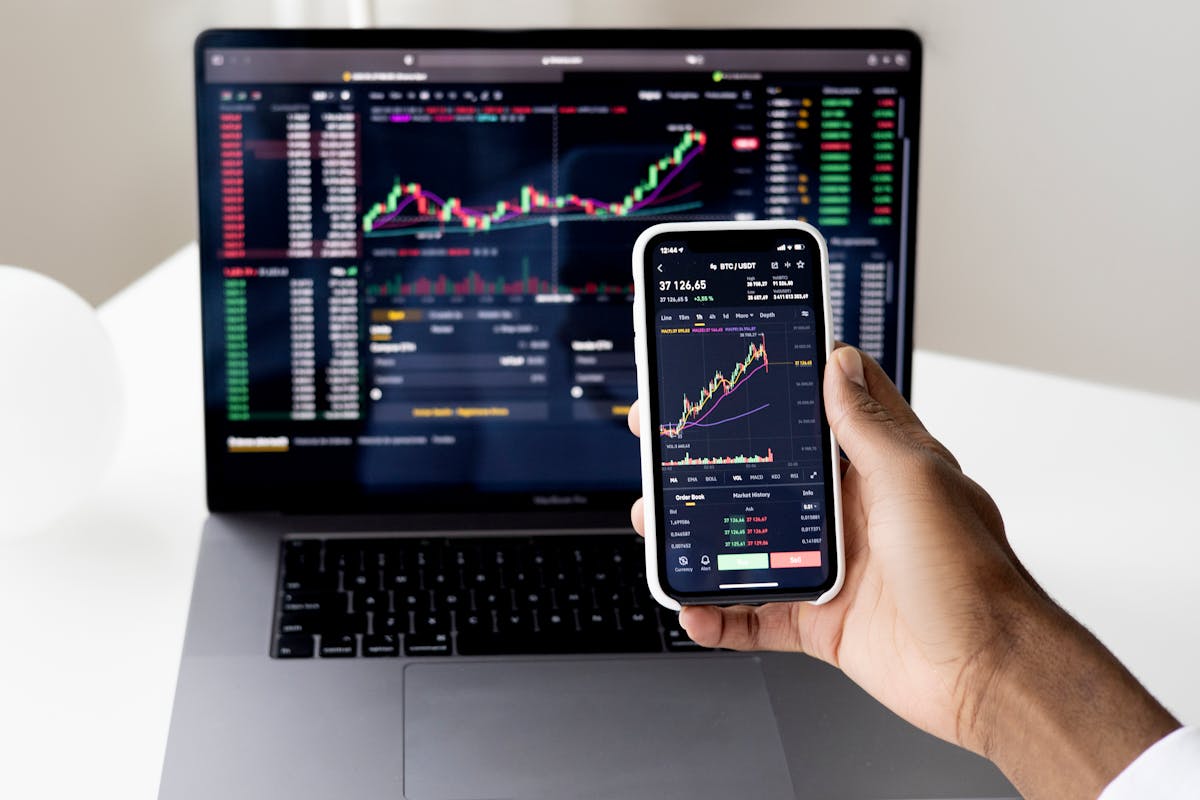 Person holding smartphone with stock market data in front of laptop displaying financial charts.