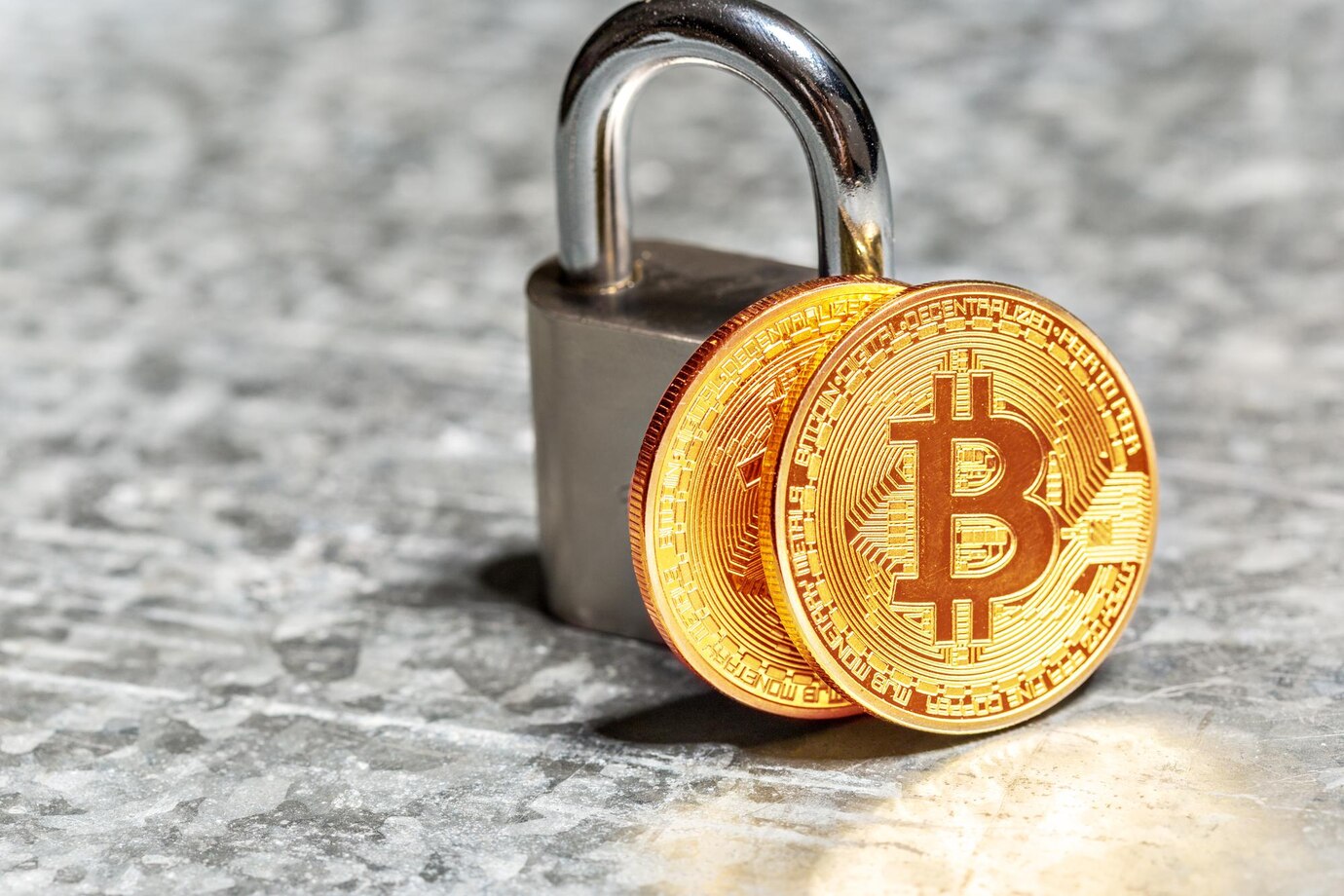 A padlock beside two gold Bitcoin coins resting on a textured gray surface, symbolizing cryptocurrency security.