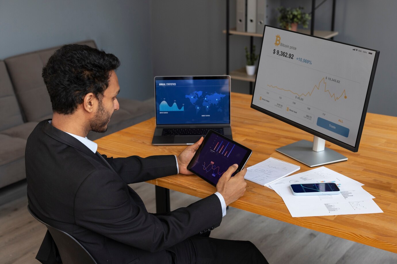 A man in a suit analyzes financial data on a tablet, laptop, and monitor in a modern office setting with papers and a smartphone on the table.