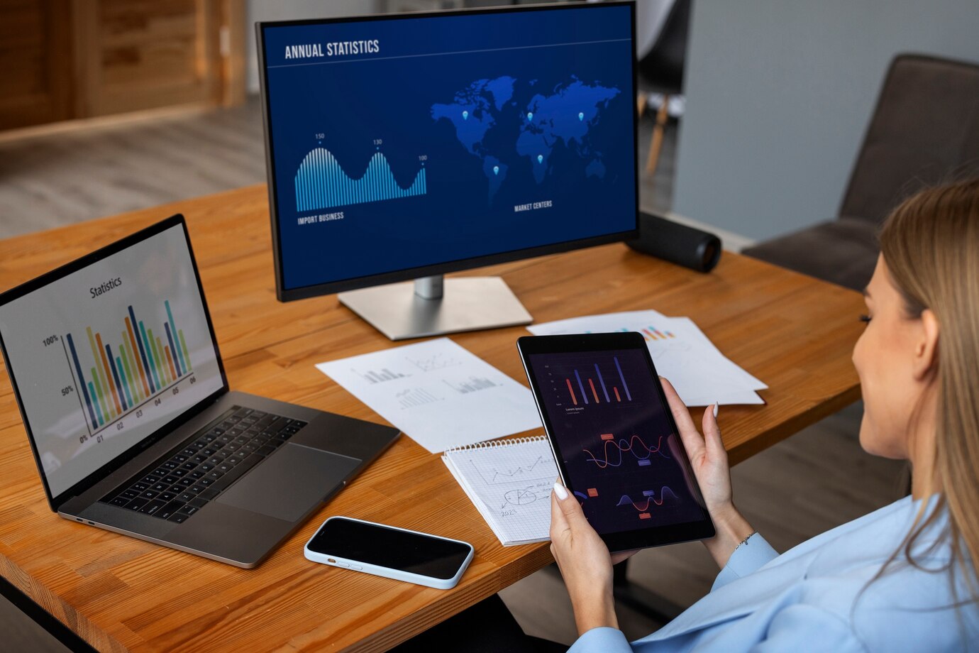 A person analyzes data on a tablet, surrounded by a laptop showing colorful statistics and paperwork on a wooden desk.