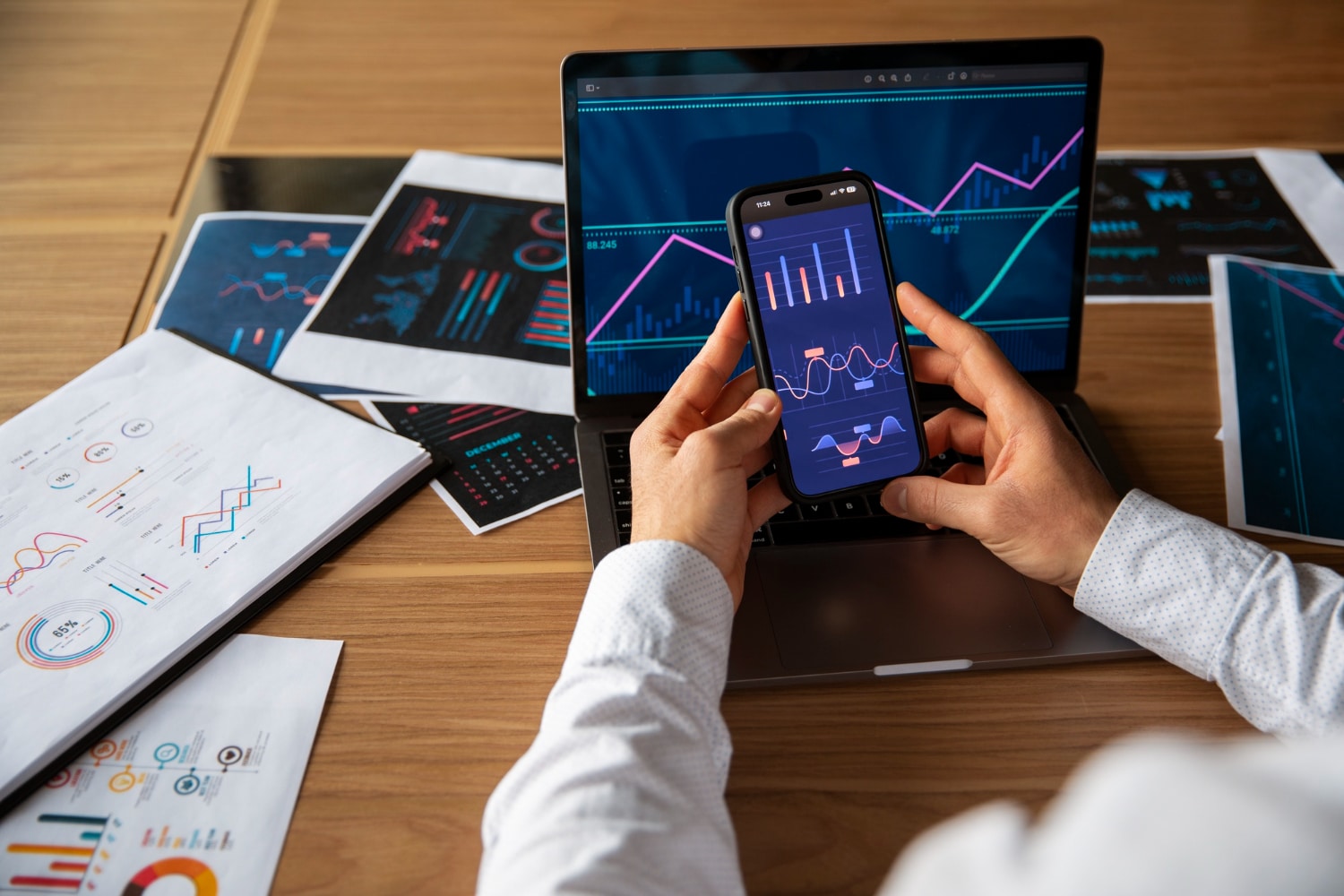 A person holding a smartphone displaying graphs, while viewing similar data on a laptop, surrounded by printed charts on a wooden table.